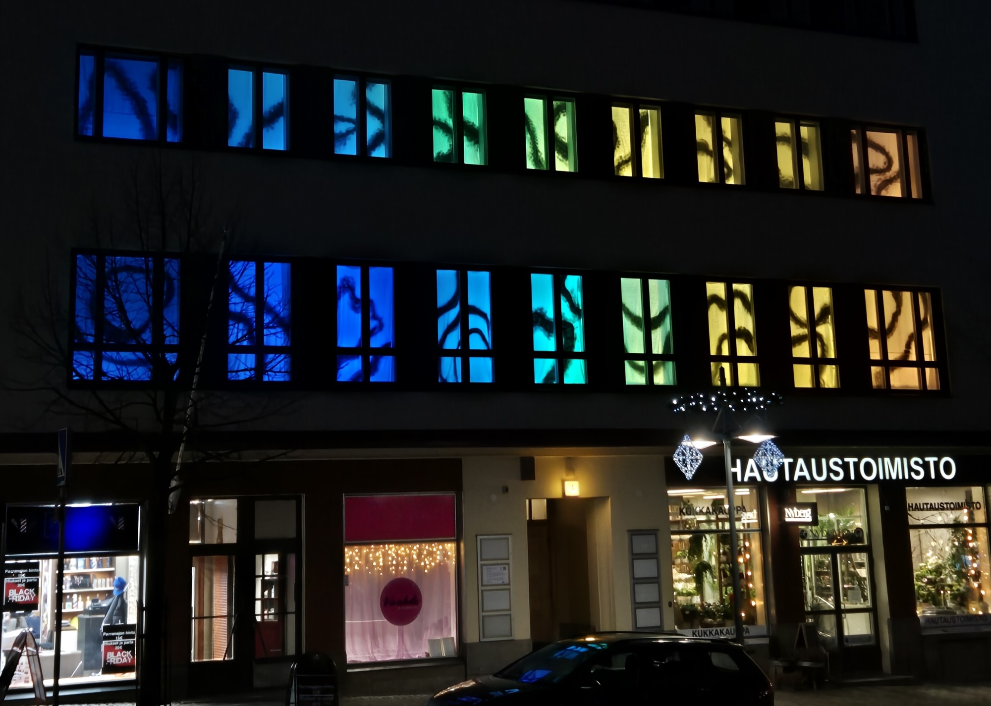 Erivärisin valoin valaistuja kerrostalon ikkunoita, katutasossa liiketiloja ja autoja parkissa.